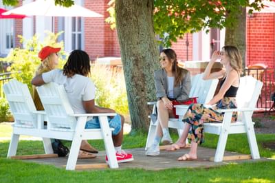 Four friends talking in white chairs under a large shade tree near patio umbrellas at Rodd Charlottetown Hotel