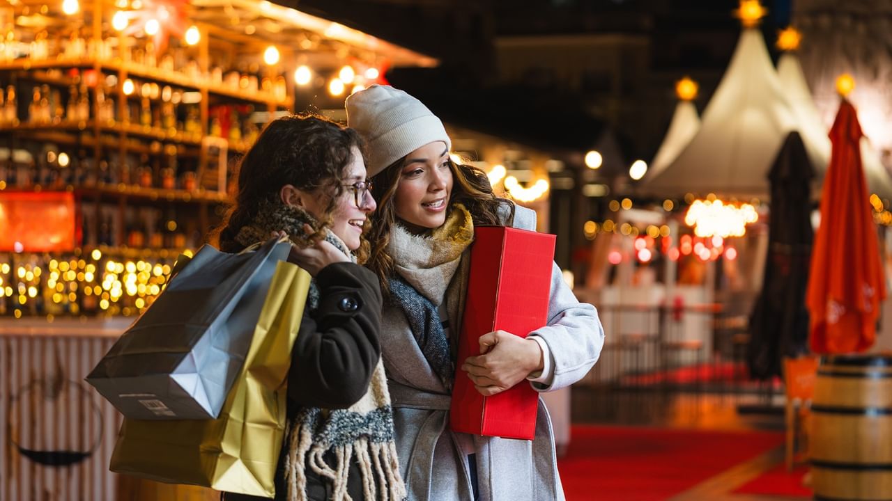 Two women carrying shopping bags and gifts at a Christmas market with festive lights and decorations.