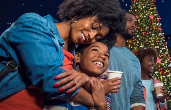 A woman holding a cup of coffee embraces a smiling boy while a man and girl look on in front of a Christmas tree. 