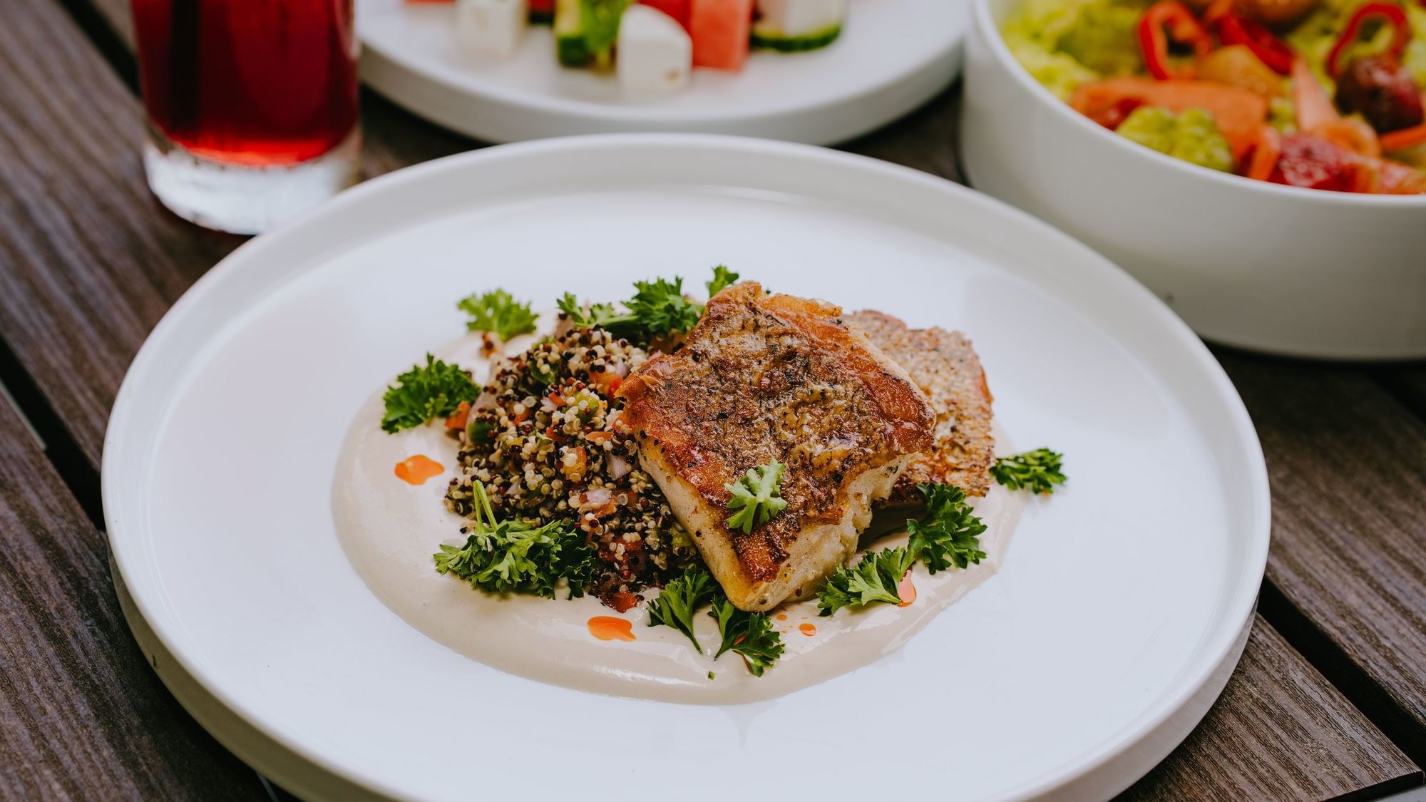 Plate of fish, quinoa, and parsley with a salad, watermelon, feta, and a red beverage on a table.