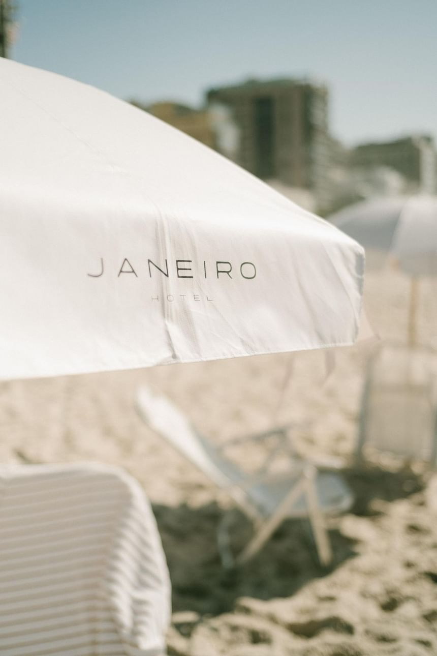 Imagem na praia do leblon de um guarda-sol branco do Janeiro Hotel e cadeiras de praia sobre a areia
