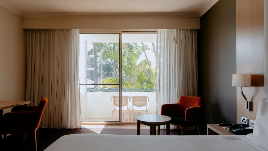 Hotel room with a bed, desk, chairs, and balcony overlooking trees.