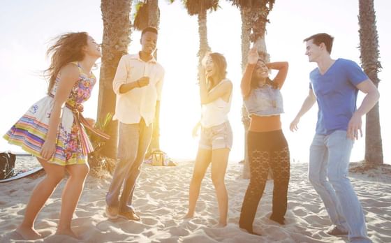 Group of friends dancing on a sunny beach, surrounded by tall palms and golden light near Hotel Angeleno
