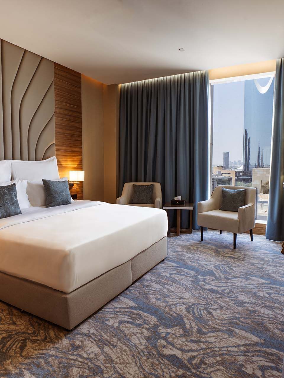 King bed with a tall fluted headboard by a window under blue drapes in Royal Suite at Cantonal Hotel by Warwick Riyadh