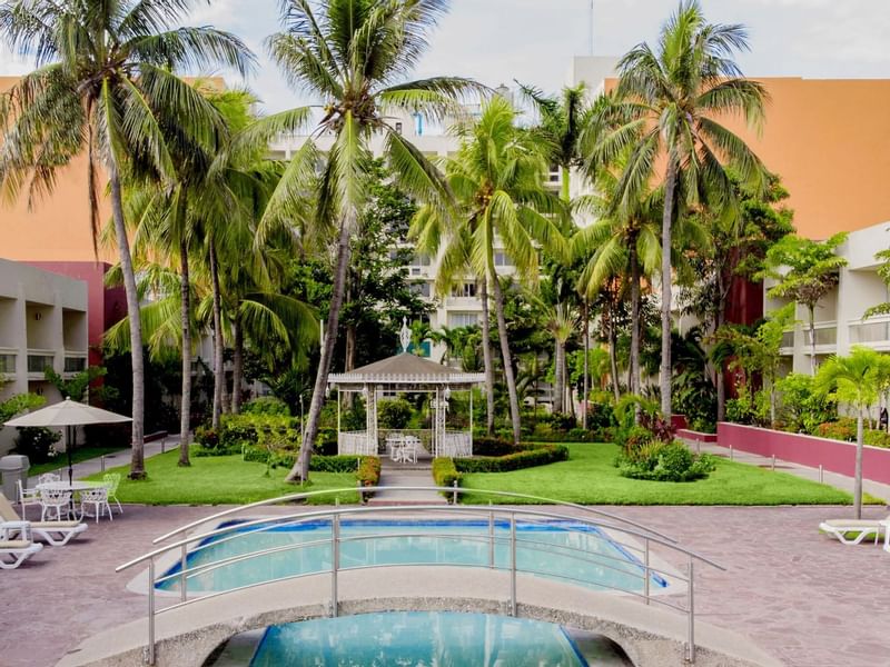 Tropical hotel courtyard at Gamma Hotels with a pool, bridge, and palm tress surrounded by lush greenery