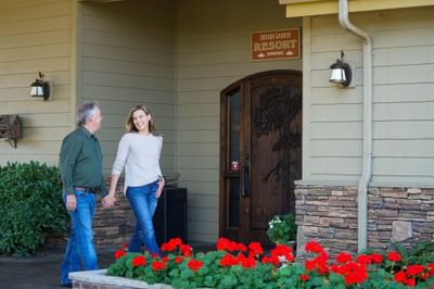 Man and woman holding hands in front of Oregon Garden Resort entrance with red flowers and stone wall.