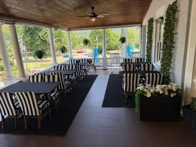Striped chairs at black tables by white flower planters, under a wood porch ceiling near a fan at Simsbury 1820 House