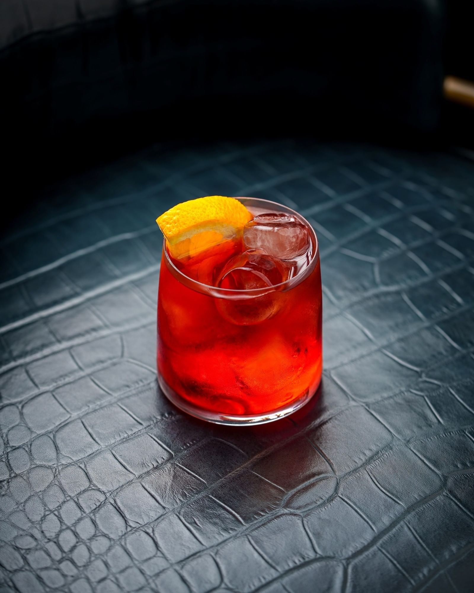 Red cocktail with ice cubes and an orange slice on textured black leather surface at Tom Brown at The Capital