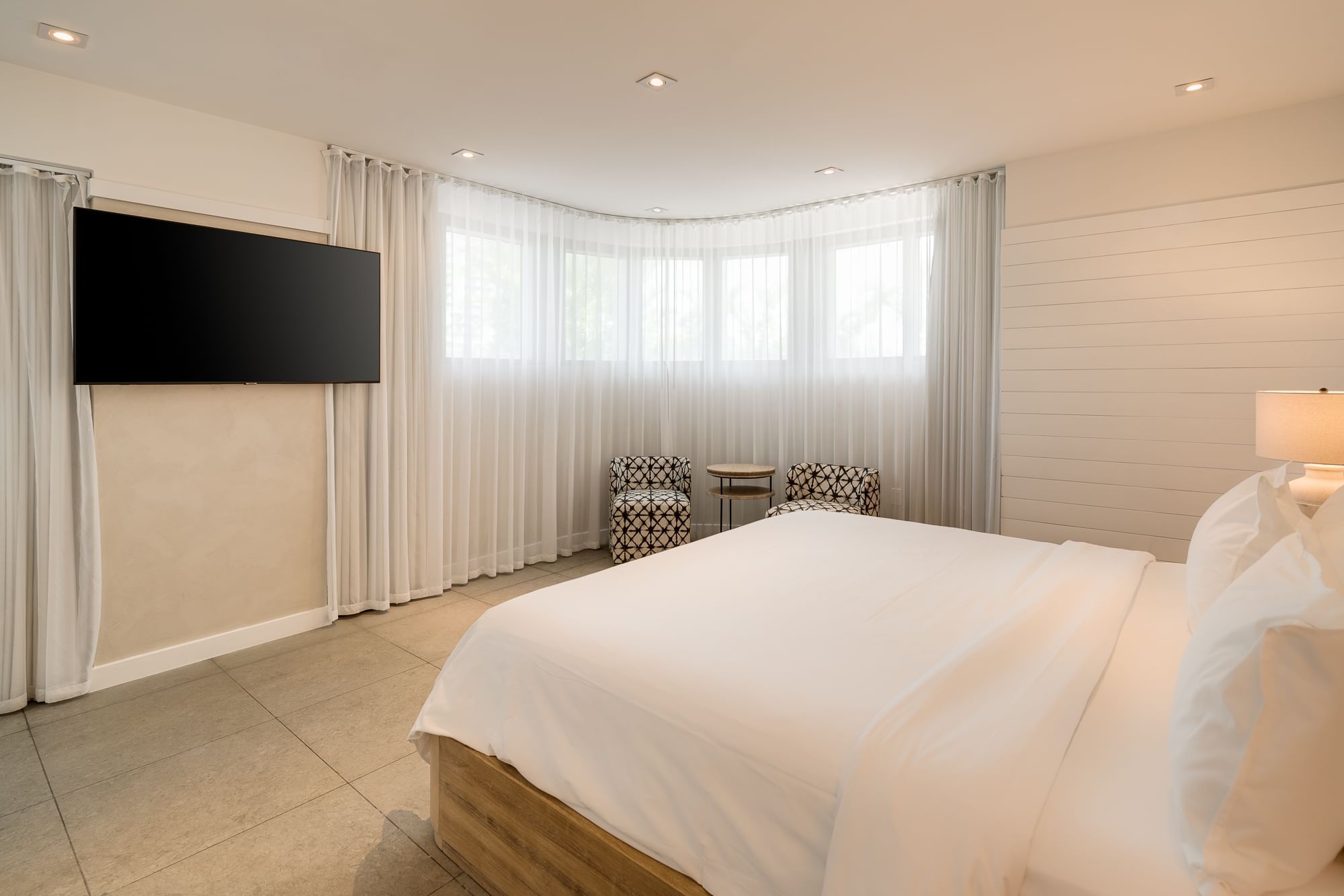 A bright and airy hotel room with a king-sized bed, flat-screen TV, and window seating area.