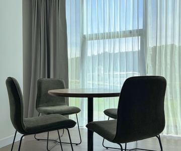Round wooden table with four dark chairs set against a sheer curtained window in the guest room at Unika Hotels
