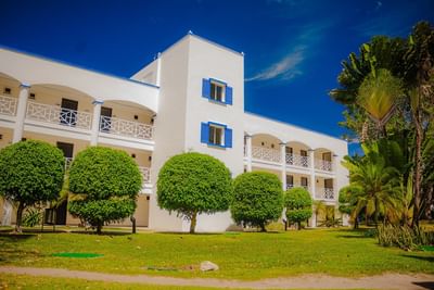 White resort building with blue shutters and balconies surrounded by manicured trees and tropical palms at Playa Blanca Beach Resort