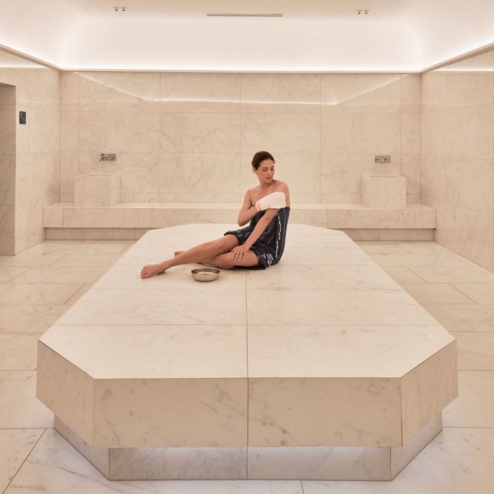 Woman in robe sits on marble bench in a tiled room, holding towel against her arm.