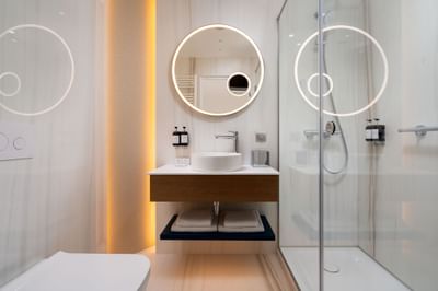 Bathroom featuring a sleek shower, minimalist vanity, round mirror, and warm lighting at Ana Hotels Europa Eforie Nord