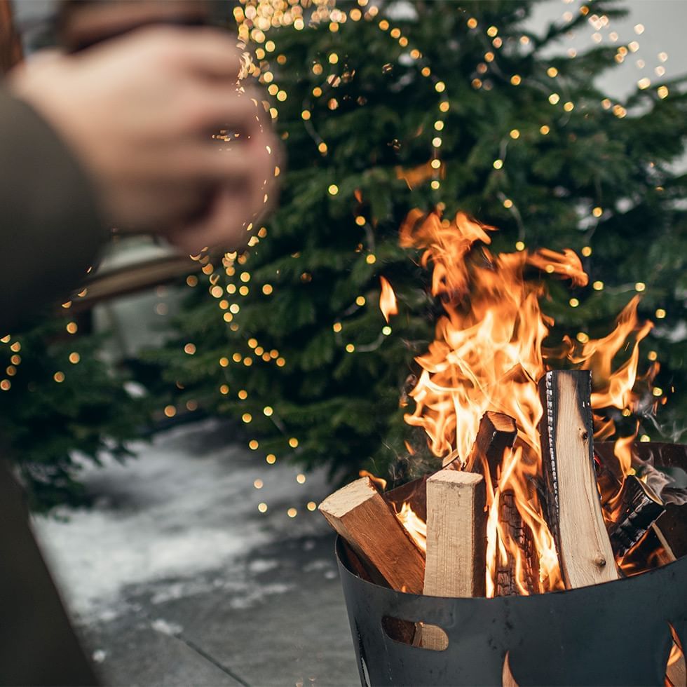 A hand sprinkles snow near a burning fire pit with logs, beside a lit Christmas tree.