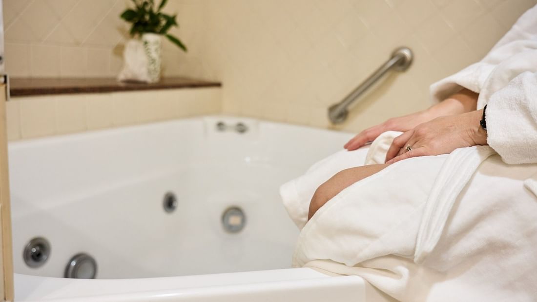 Woman in a bathrobe sitting on the edge of a white bathtub with a plant on the side.