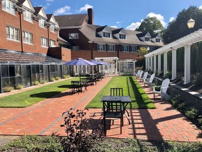 Brick patio courtyard with dining tables under purple umbrellas facing green lawn and pergola at The Simsbury Inn