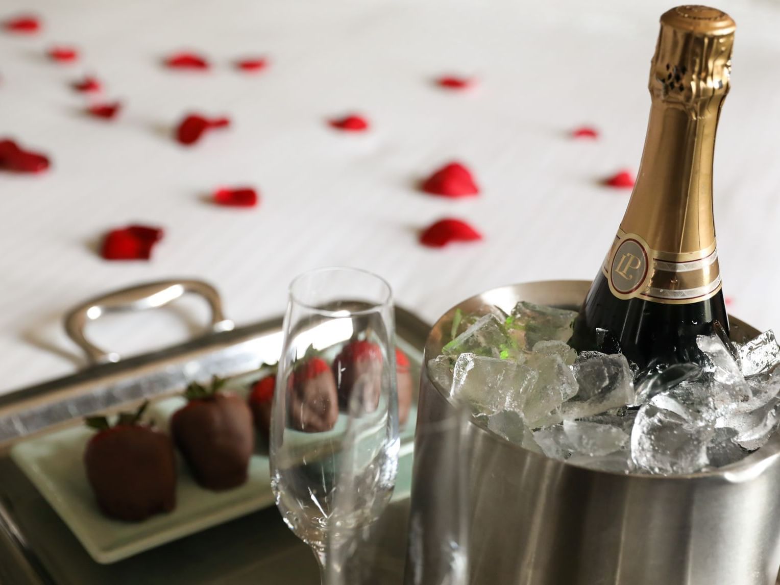 A bucket of champagne and chocolates on a white bed scattered with red rose petals