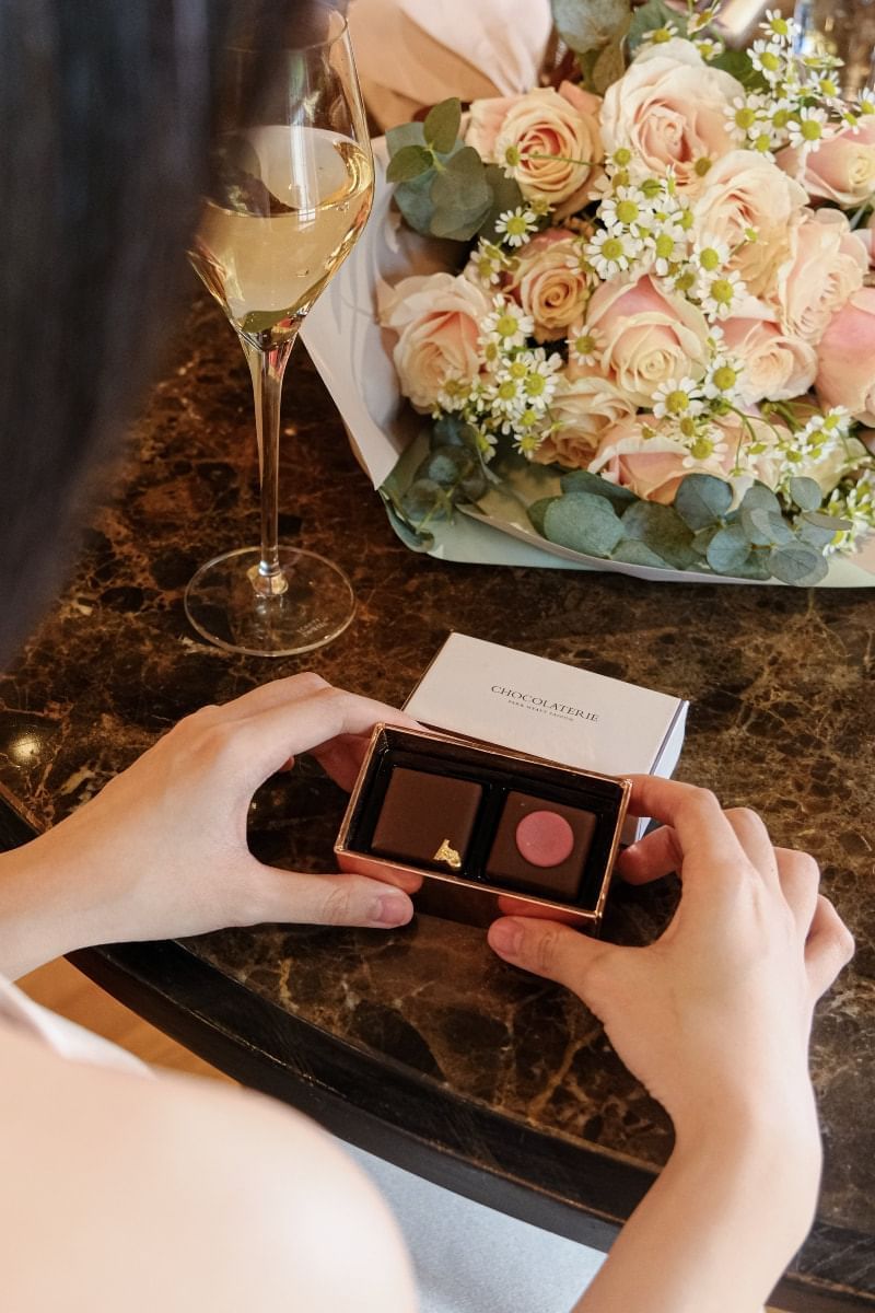 Person holding a small box of chocolates beside a bouquet of pink roses and a glass of champagne at Park Hyatt Saigon