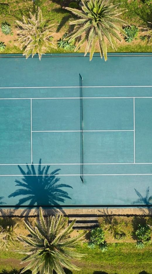 Drone view of the outdoor tennis court at Caloura Hotel Resort, bordered by lush green palm trees casting shadows