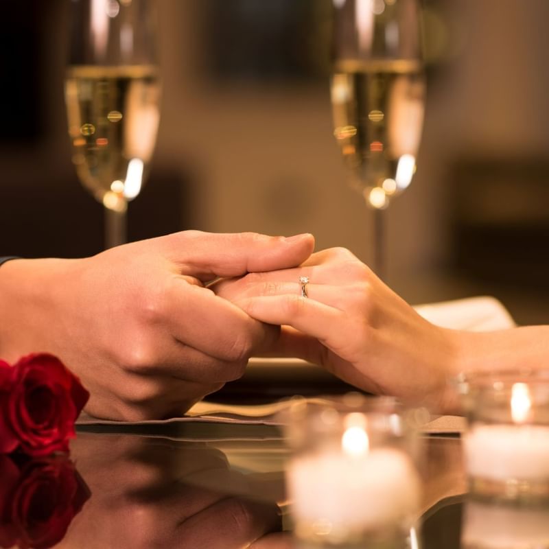 A man and woman holding hands, with a rose and champagne glasses on a table at Warwick Denver