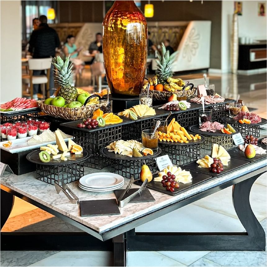 Bufé de Bazaar con fruta, queso y carnes bajo un jarrón alto sobre una mesa de mármol en Megapolis Hotel Panama
