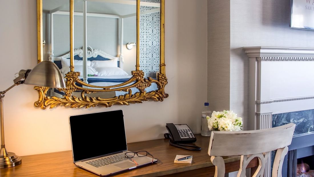 Laptop on a wooden desk by a gold mirror & white flowers under a grey wall in Loft Fireplace Suite at Warwick San Francisco