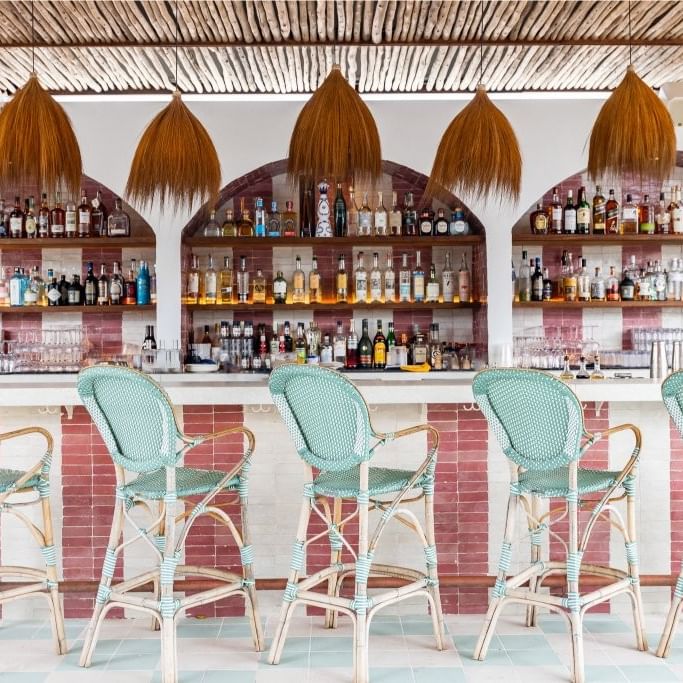 Tropical bar with thatched pendant lights, turquoise woven barstools, and shelves of spirits for complimentary guest drinks.