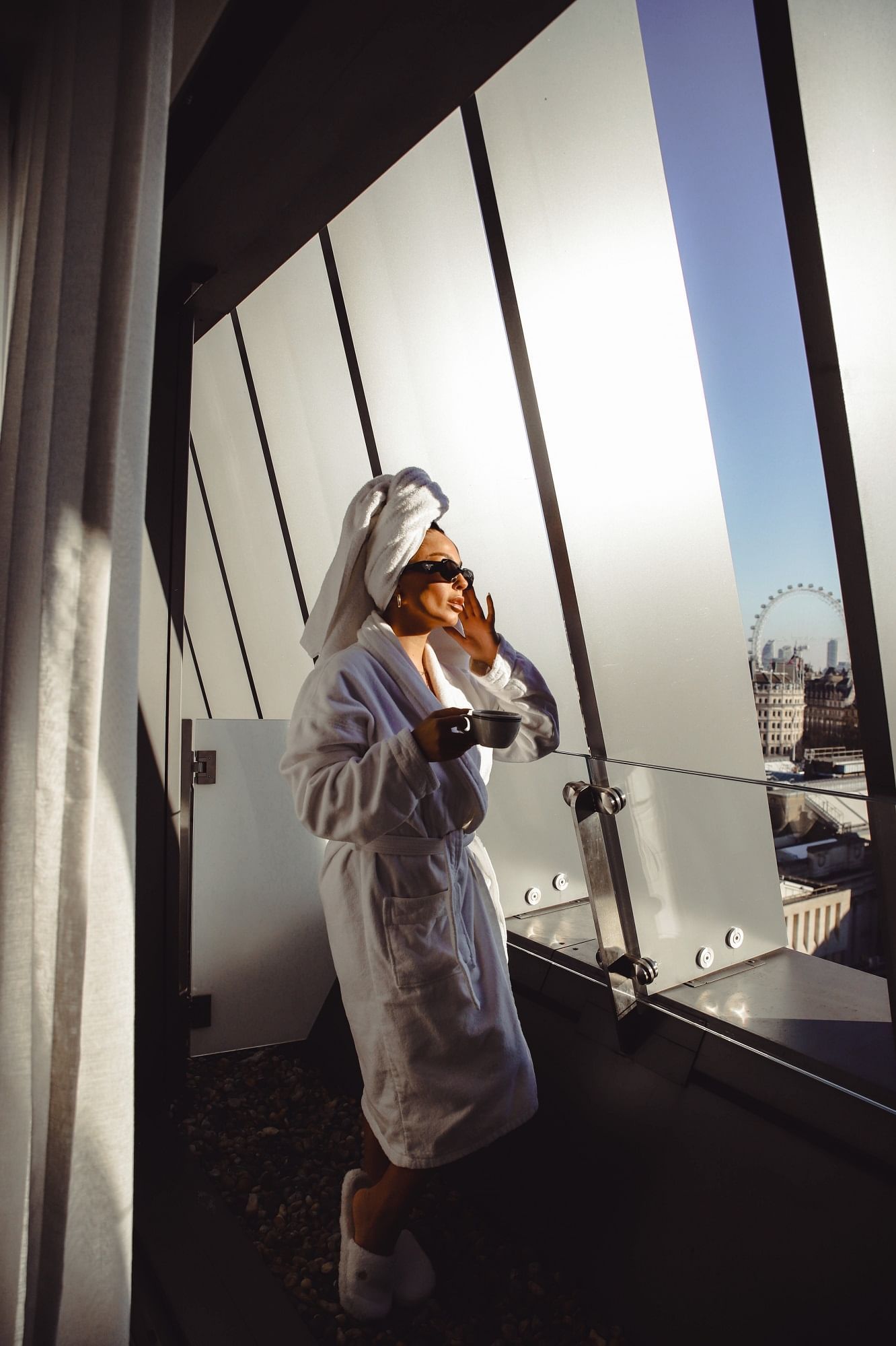 A woman in a bathrobe standing by the balcony overlooking the cityscape while having tea at The Londoner