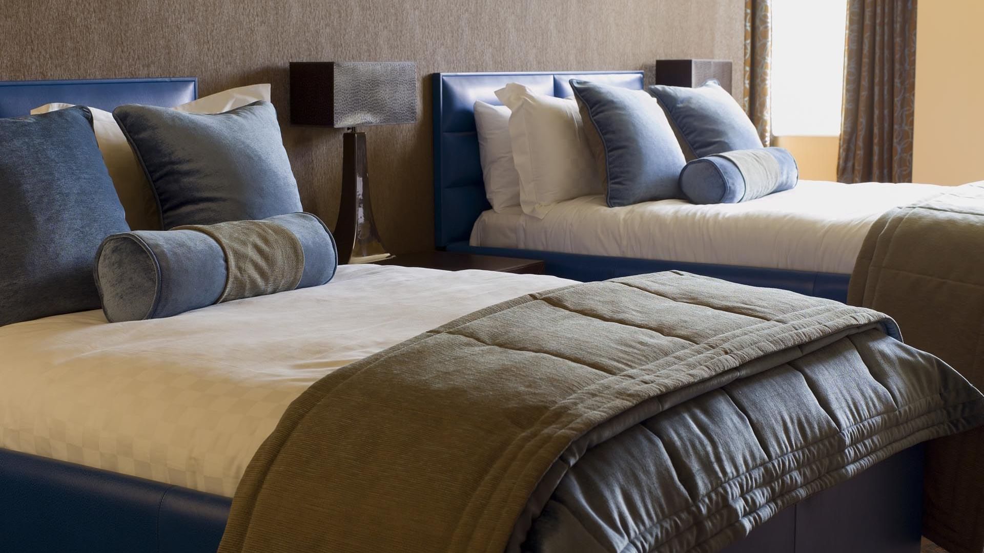 Two neatly made beds with blue padded headboards and bolsters in The Azure Suite at The May Fair Hotel, London