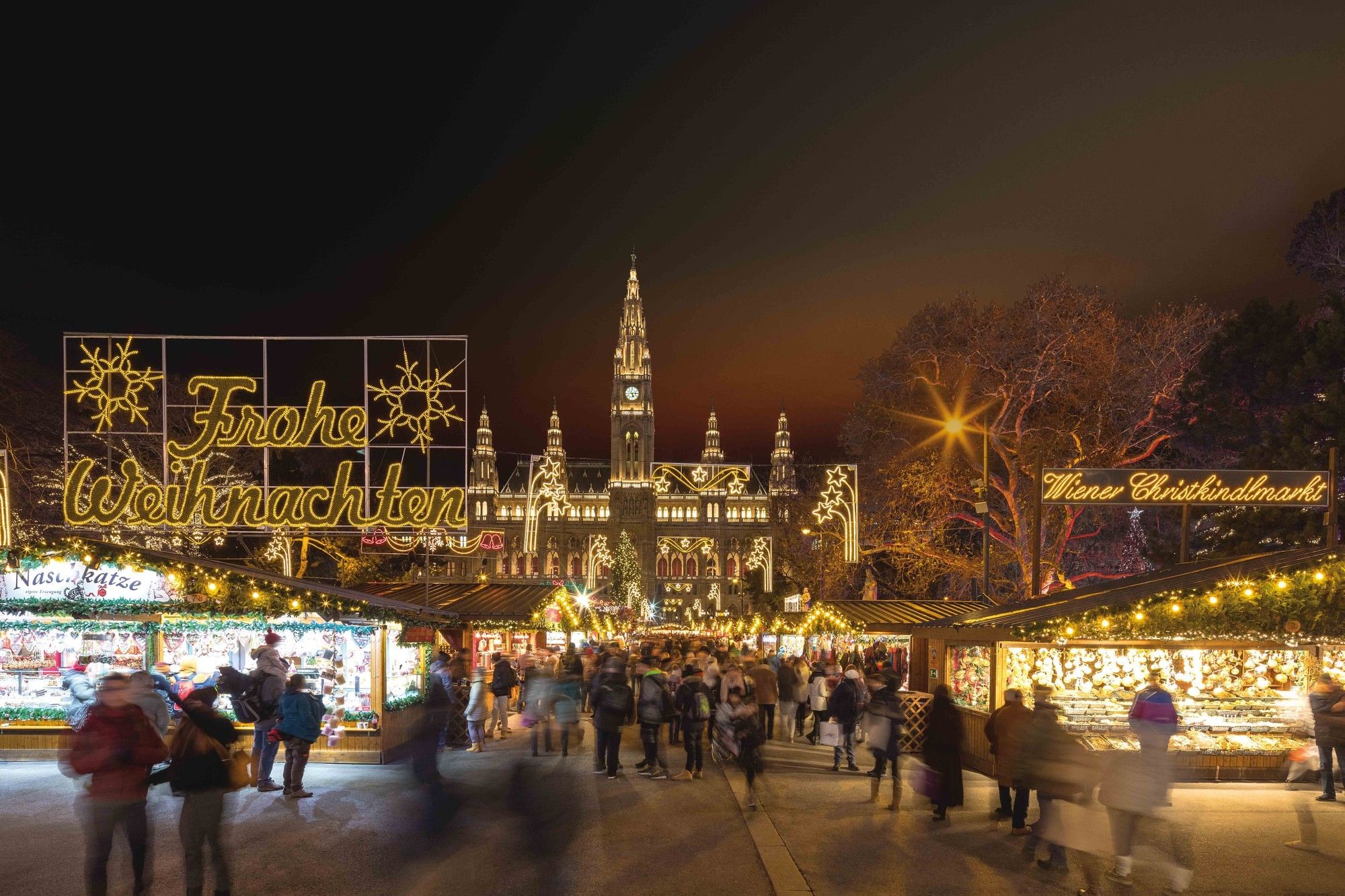 People stroll through the festively lit Vienna Christmas Market in front of City Hall, with a “Merry Christmas” sign in the foreground.