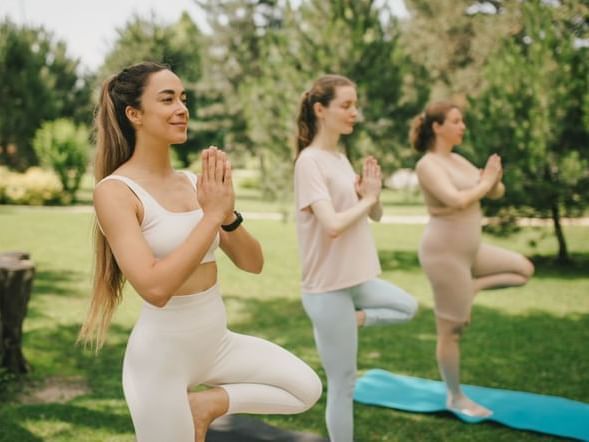 Three women practicing yoga in tree pose with hands in prayer, in a serene park at Fiesta Americana Travelty