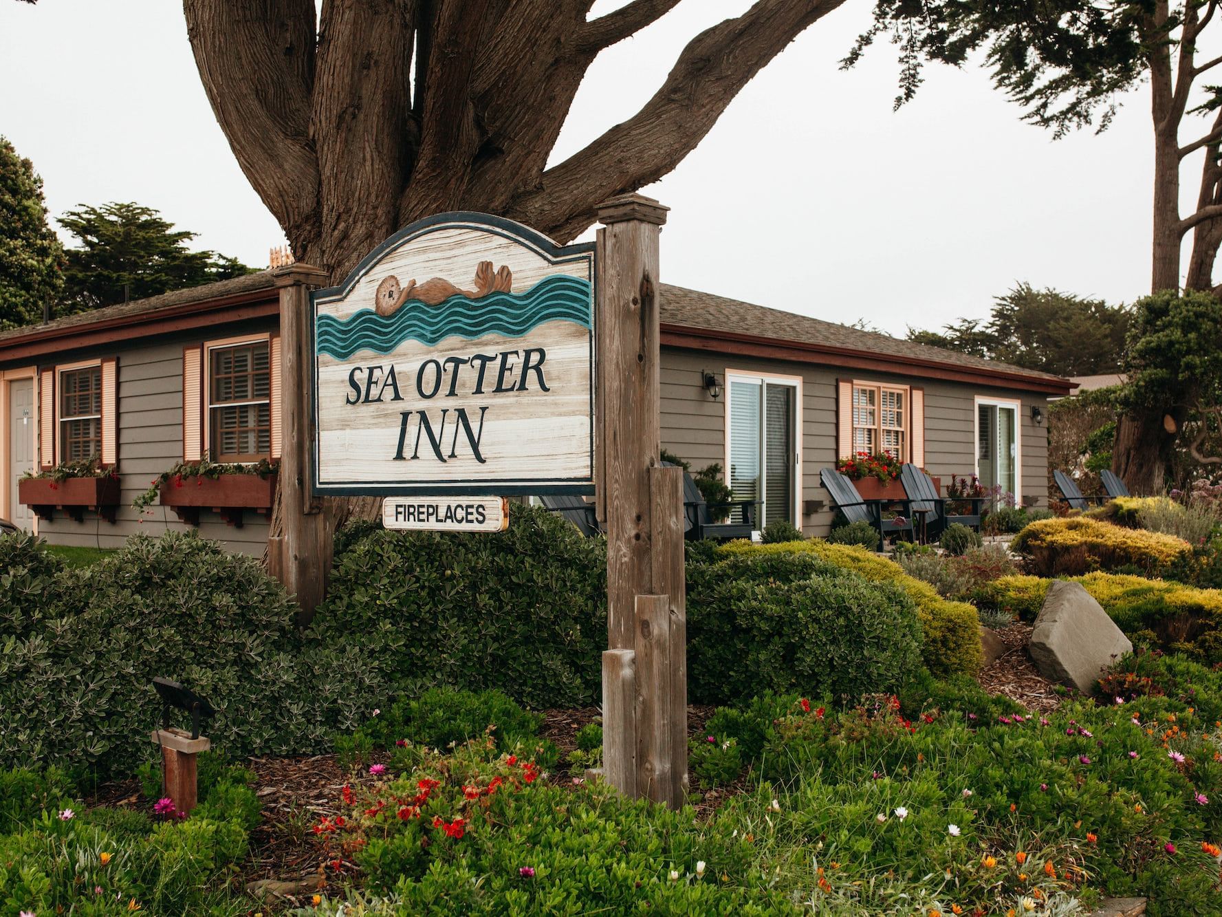 Wooden sign by lush bushes under a tall tree, surrounding the exterior guest rooms at Sea Otter Inn
