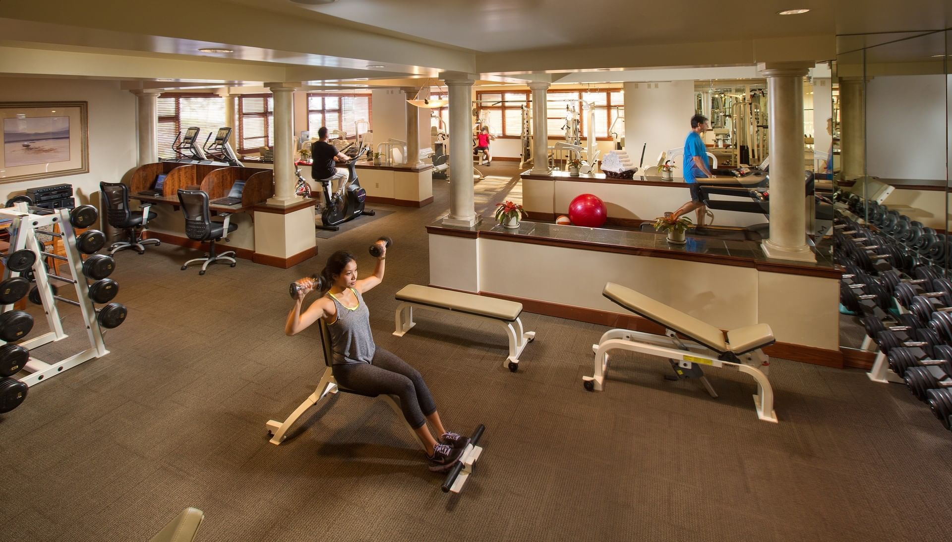 A well-equipped gym with a woman exercising on a bench press, surrounded by various exercise equipment, mirrors, and columns.