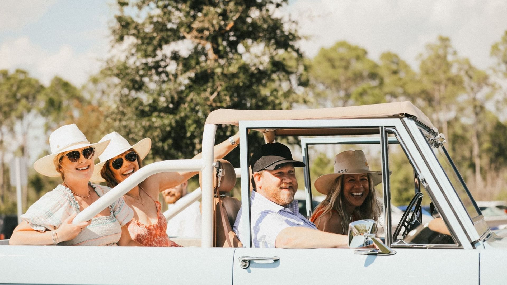 Group of smiling friends wearing sunglasses and hats riding in a white vintage convertible on a sunny day outdoors.