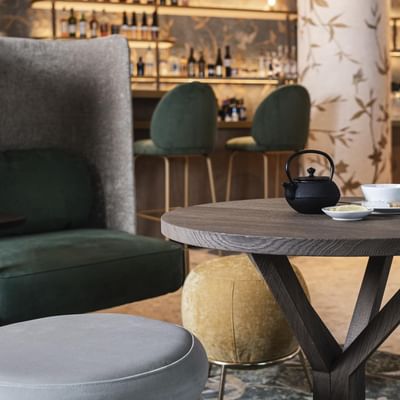 Tavolo circolare con teiera e tazza, affiancato da poltrone e sgabelli in un bar.