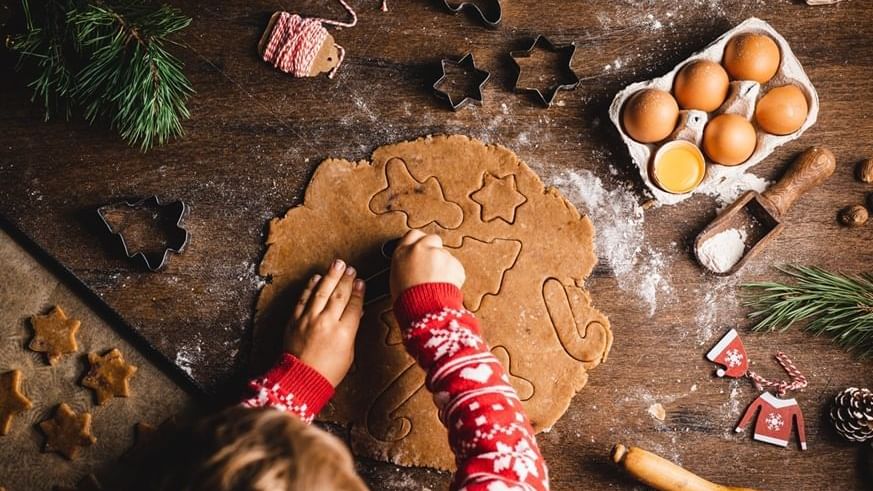 Child cutting out cookies with holiday-themed cutters and festive decor around.