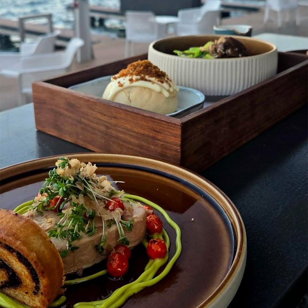 Two dishes on a table: a plate with meat, vegetables, and bread, and a wooden tray with desserts.