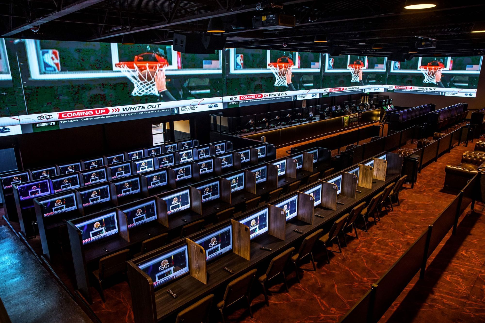 Rows of betting screens and chairs under basketball displays, beside a long counter and seating at The Brook