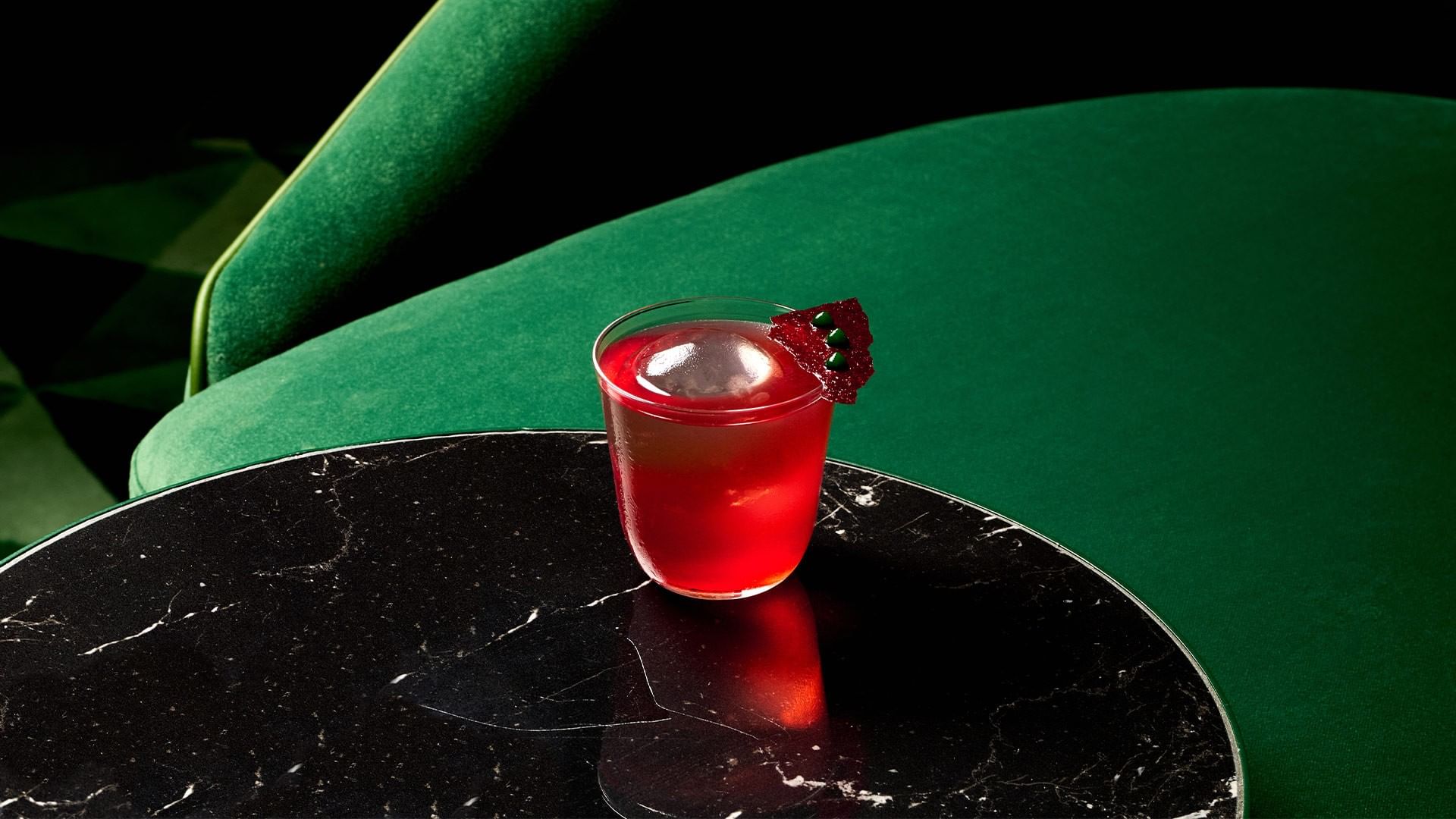 A red cocktail with garnish on a black marble table next to a green chair.