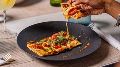Cheese-pull flatbread slice held above a tomato and microgreen-topped pizza on a black plate at Sugar Cane Club Hotel & Spa