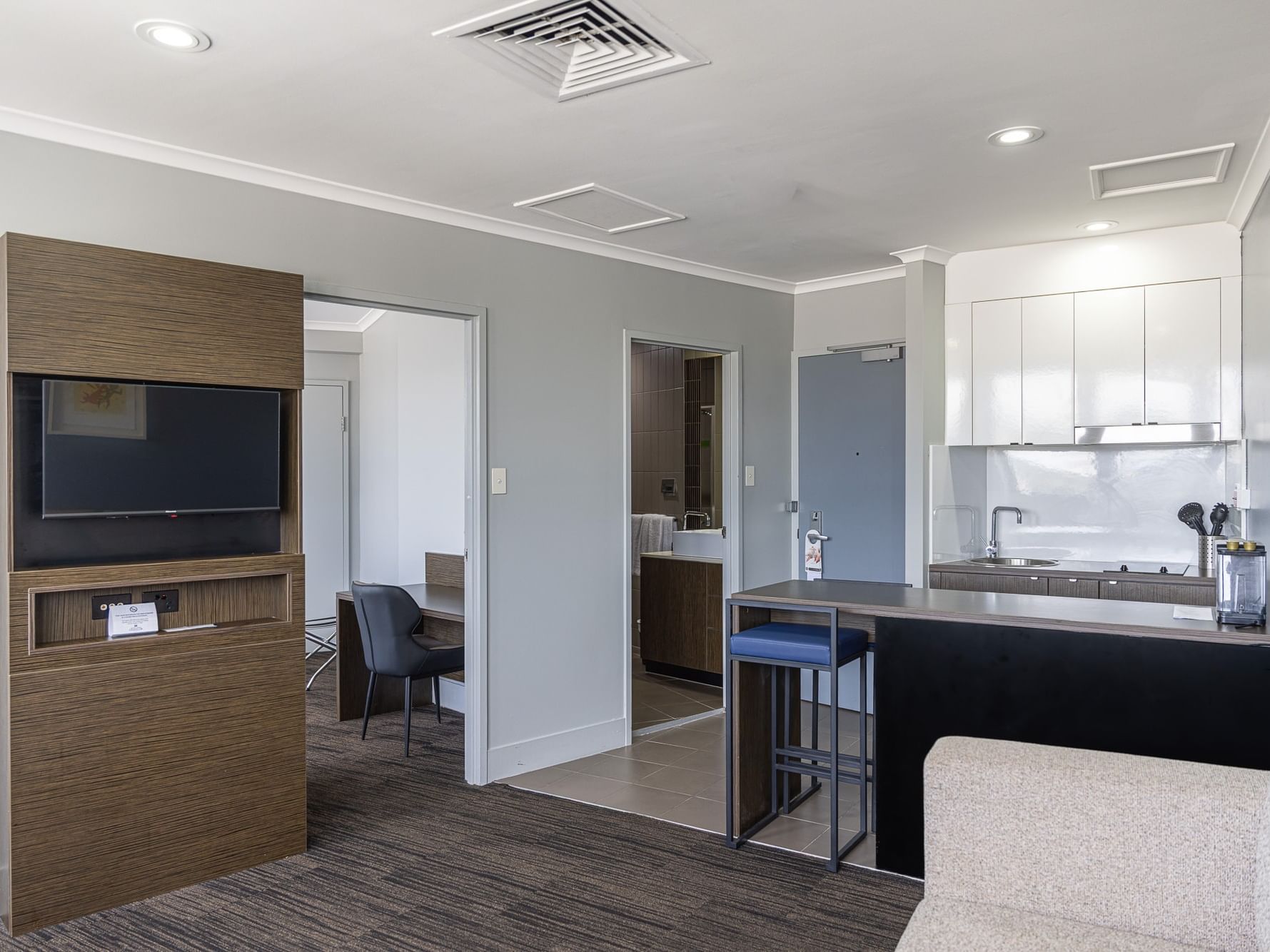 Modern hotel suite featuring a living area with a wall-mounted TV, a small kitchenette, a breakfast bar with a blue stool, and a view into the bedroom.