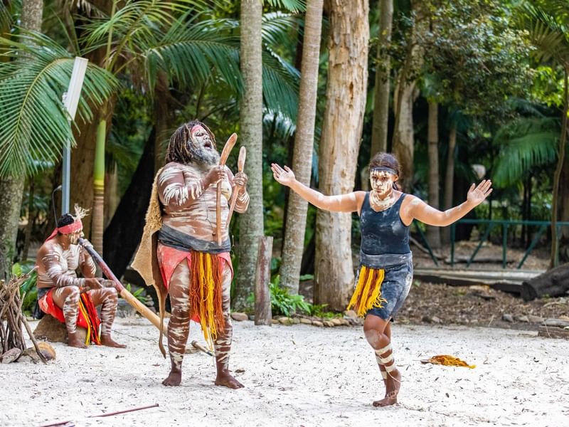 Three First Nations people in traditional dress and body paint performing on a beach near Sofitel Brisbane Central