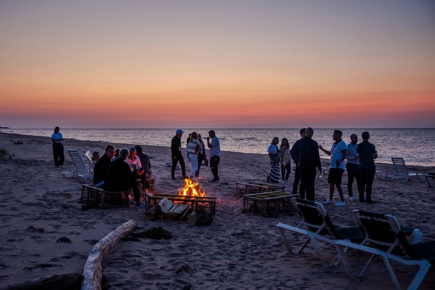 Guests gathering around a beach bonfire on the sand during a vibrant sunset at Rodd Crowbush Golf & Beach Resort