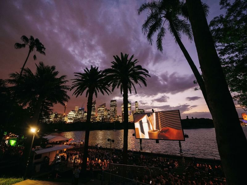 Crowd watching Westpac OpenAir Cinema outdoors at night with palm trees and city skyline in background.