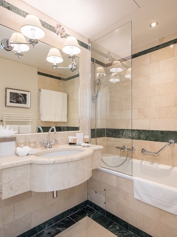 Marble vanity by a glass-enclosed bathtub under warm lights with rolled towels in Classic Single Room at Warwick Geneva