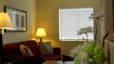 Cozy living room with burgundy sofa, table lamp, white flowers, and window blinds at London Bridge Resort
