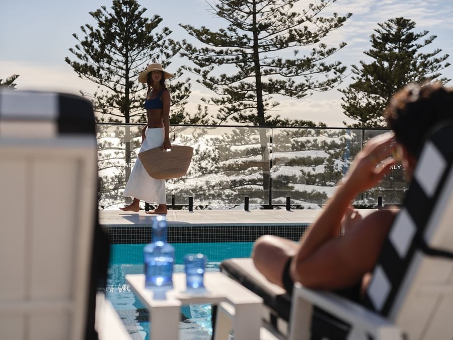 Woman walking by the swimming pool at Elysium Manly, with pine trees under a clear sky and beach chairs near glass fences