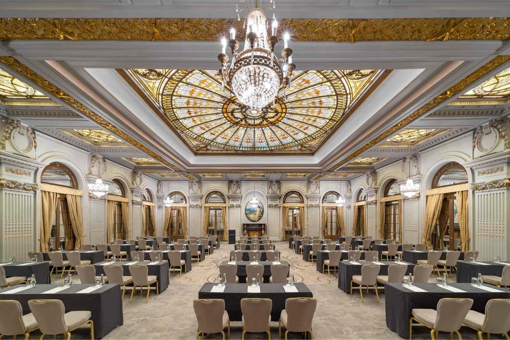 Sala Le Diplomate de la InterContinental Athénée Palace Bucharest, cu candelabre, plafon decorativ din sticlă și mese pregătite pentru un eveniment.