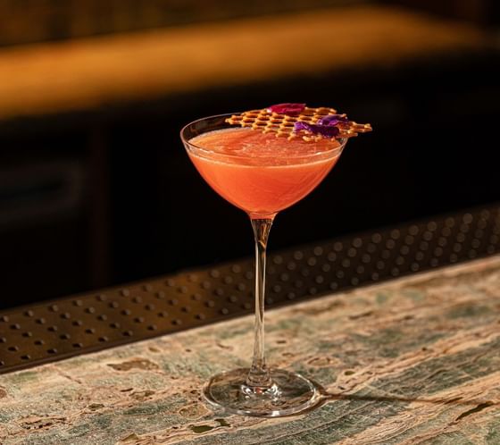 A pink cocktail garnished with a golden lattice on a textured green bar surface at The Kitchens