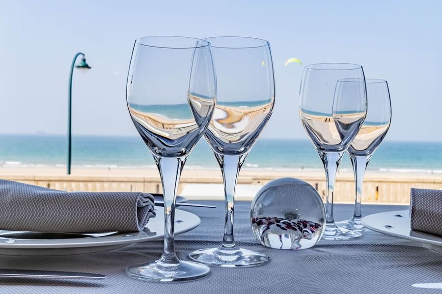 Wine glasses arranged on a white tablecloth with a sunny beach view in the back at Le Grand Hôtel des Thermes